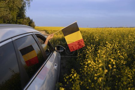 Europareise mit dem Auto. Das Auto fährt an einem sonnigen Tag offroad durch ein blühendes gelbes Rapsfeld. Eine deutsche Fahne ragt aus dem Fenster. Nationales Symbol für Freiheit und Unabhängigkeit