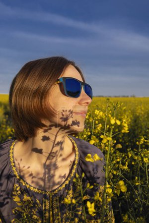 Sensual female portrait against the backdrop of a blooming yellow rapeseed field and blue sky, shadows on the face from flowers. A woman in blue sunglasses and a Ukrainian embroidered blouse