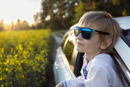 7-8 Jahre alter Junge mit blauer Sonnenbrille blickt aus dem offenen Autofenster und betrachtet interessiert die Natur. Reisen mit der Familie im Sommer