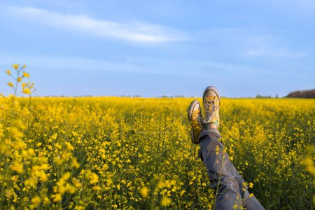 Frauenbeine in gelben stylischen Turnschuhen vor dem Hintergrund eines blühenden gelben Rapsfeldes und blauen Himmels. Spaziergang, Harmonie, Energie der Natur, Entspannung, Aktivität, Stimmung, Freiheit