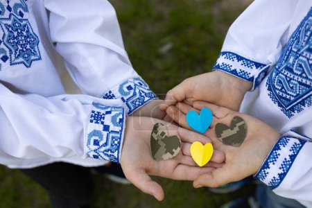4 hearts cut from military camouflage fabric and yellow-blue paper lie on palm of two children in Ukrainian embroidered shirts. Stop war in Ukraine. With love and gratitude to soldiers of Armed Forces