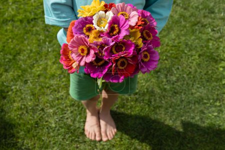 bouquet of bright flowers in the hands of a barefoot child standing on the lawn on a sunny summer day. Flower gift for mom or grandma. joy, cheerful positive atmosphere. Floral surprise with love
