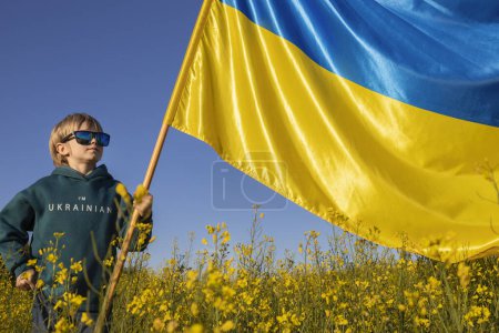 Junge in Kapuzenpulli mit Aufschrift Ich bin Ukrainer mit großer gelb-blauer Flagge vor einem Rapsfeld. Das ukrainische Kind wünscht seinem Heimatland Frieden. Nationales Symbol für Freiheit und Unabhängigkeit