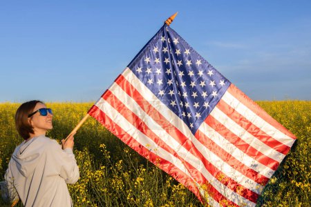 woman holds a large American flag against background of blue sky and rapeseed field on sunny day. holiday. Independence Day of the United States of America. Pride, freedom. traveling around country