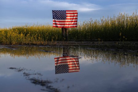 American flag and its reflection in the water against the background of a blooming yellow rapeseed field and the sky. Independence Day. National symbol of freedom of the States of America