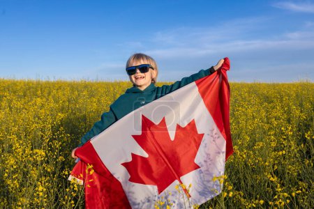 Canadian flag in the hands of a joyful boy against the backdrop of a blooming rapeseed field. Canadian Independence Day. Pride, freedom, patriotism. Travel around the country. National symbol