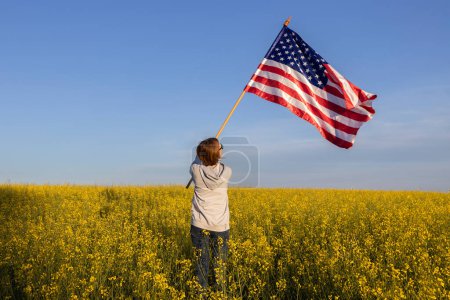 Die amerikanische Flagge liegt in den Händen einer Frau, die mit dem Rücken zwischen blühenden gelben Rapsfeldern steht. US-Unabhängigkeitstag am 4. Juli. Stolz auf die eigene Heimat, Freiheit, Patriotismus, Demokratie. Reisen durch das Land