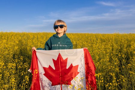 niño vistiendo sudadera con capucha con inscripción Soy ucraniano, de pie entre campo de colza amarilla con bandera canadiense. Agradecimiento a Canadá por parte del pueblo ucraniano por su ayuda y apoyo. Diáspora.