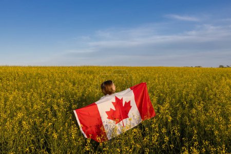 Frau steht mit dem Rücken inmitten eines gelben, blühenden Rapsfeldes und hält an einem sonnigen Tag stolz eine große kanadische Flagge in der Hand. Tag der kanadischen Unabhängigkeit. Freiheit, Vertrauen, Stolz, Nationalsymbol