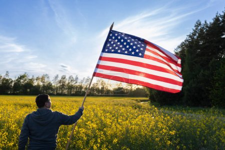 Mann steht mit dem Rücken und hält an einem sonnigen Tag eine große amerikanische Flagge vor blauem Himmel und Rapsfeld. Urlaub. Unabhängigkeitstag der Vereinigten Staaten von Amerika. Reisen durch das Land