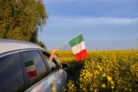 Europareise mit dem Auto. Eine italienische Flagge ragt aus dem Fenster. Nationales Symbol für Freiheit und Unabhängigkeit. Ein Auto fährt an einem sonnigen Tag im Gelände durch ein wunderschön blühendes gelbes Rapsfeld
