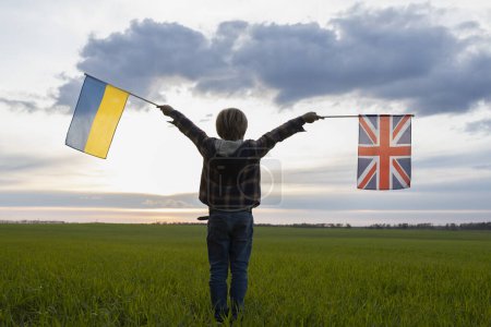 niño irreconocible sostiene banderas británicas y ucranianas contra el cielo del atardecer. banderas de Ucrania y Gran Bretaña. Concepto de cooperación y asociación entre dos países europeos.