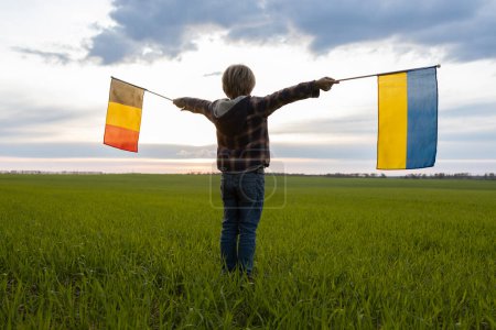 unrecognizable child holds German and Ukrainian flags against the background of the sunset sky. flags of Ukraine and Germany. Concept of cooperation and partnership between two European countries
