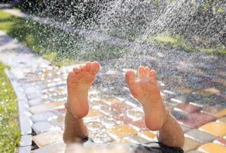 Das Kind liegt barfuß auf einer Pflasterplatte unter Wasserspritzern aus einem Schlauch oder Brunnen. Kinderfüße in der Sonne und im Wasser. Sommerspaß. Frohsinn. Spaß haben. Nahaufnahme