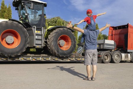 man and boy sitting on his shoulders stand in front of large tractor loaded on truck trailer. son's passion for cars. Father's Day, be together. child wants to become driver or mechanic, like dad.