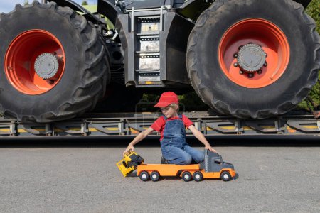 boy plays with toy cars. Behind him stands a real tractor with huge wheels. The child wants to become a driver or mechanic, like dad. commercial vehicles and business equipment