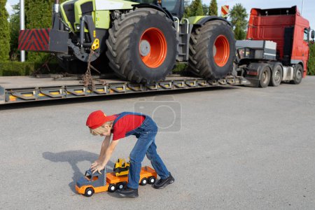 Junge spielt mit Spielzeugautos auf dem Parkplatz neben einem richtig großen Lastwagen mit beladenem Traktor. Das Kind träumt davon, wie Papa Fahrer oder Mechaniker zu werden. Nutzfahrzeuge für Unternehmen