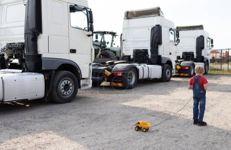 boy plays with a toy dump truck on the control panel. Behind the child there are real big trucks in a row. The boy's hobby is construction equipment and big cars. Be a driver or mechanic like dad