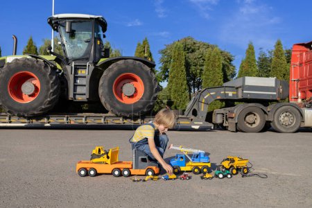 7-year-old boy plays with toy cars while a huge tractor on a transport trailer stands behind him. interesting childhood of a child, games in the profession
