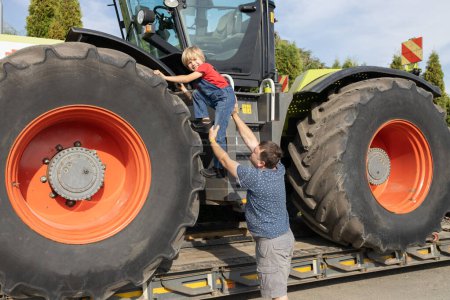 boy climbs onto a huge tractor wheel, his dad helps him. Spend the day with your son. Interest in machines and their operation. interesting childhood. Contrast sizes