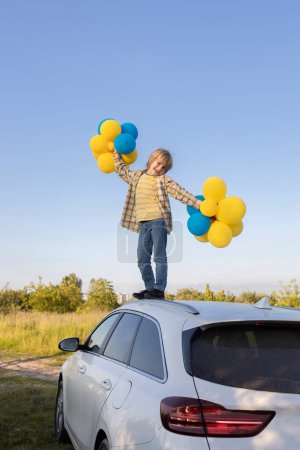 heureux garçon debout sur le toit de la voiture et tenant de nombreux ballons jaunes et bleus contre le ciel. se sent bien, vacances, anniversaire. Jour de l'indépendance de l'Ukraine. Le jour du ballon. désir de paix pour l'Ukraine