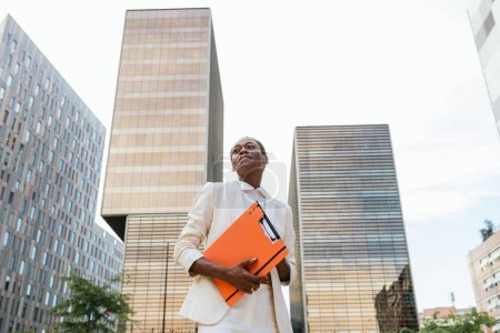 This photo features a confident African businesswoman in her fifties holding an orange folder, exuding power and authority. Her short afro hair and white business attire add to her professional look