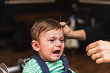 Foto de El niño llorón se corta el pelo por primera vez en la peluquería, expresando miedo e incomodidad durante el proceso. - Imagen libre de derechos