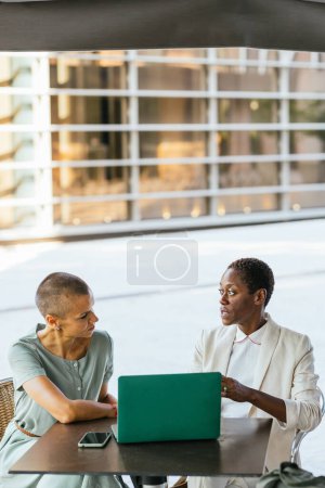 Two professional women engage in a discussion over a laptop at an outdoor setting