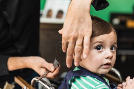 Kleinkind erlebt beim Friseur seinen ersten Haarschnitt, wobei ihn eine Friseurhand sanft führt