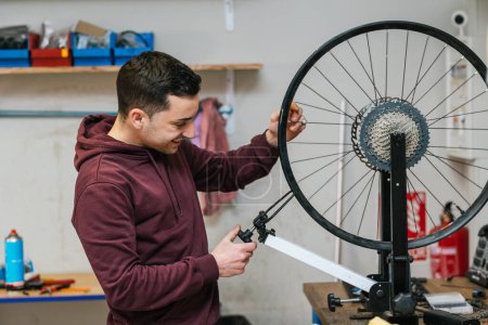 A smiling bike mechanic adjusts and repairs the rear wheel rim of a bike on the workbench in his workshop.