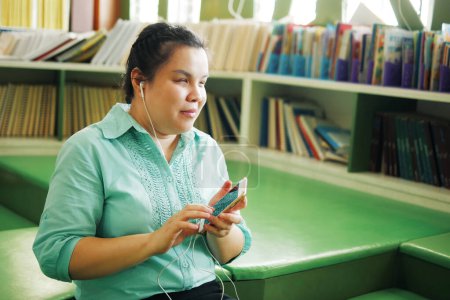 Téléchargez les photos : Portrait d'une femme asiatique atteinte de cécité portant des écouteurs utilisant un téléphone intelligent avec accessibilité vocale pour les personnes ayant une déficience visuelle dans une bibliothèque de travail créative. - en image libre de droit