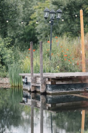 Rustikale Holzbrücke über Teich mit schönem Gartenhintergrund. Ländliche Landschaft im Sommer. Konzept für entspannende Outdoor-Aktivitäten. Rückzugsort auf dem Land mit natürlichem Pool. Grüner Lebensstil.  