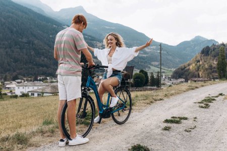 Téléchargez les photos : Deux amis ou couple font du vélo le long de la route. Sport et vie active concept de coucher de soleil. Couple apprendre à faire du vélo, s'amuser ensemble, voyager dans les montagnes près de la ville - en image libre de droit