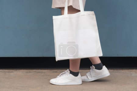 Photo for Blank white tote bag canvas fabric with handle mock up design. Close up of woman holding eco or reusable shopping bag on grunge blue metal wall. No plastic bag and ecology concept. - Royalty Free Image