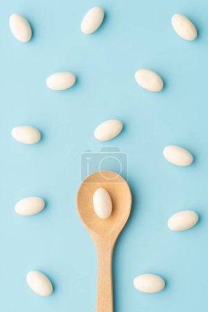 Téléchargez les photos : Capsules de gélatine molle blanche avec cuillère en bois sur fond bleu clair. Concept de supplément probiotique, vitamines et addiction aux analgésiques. - en image libre de droit