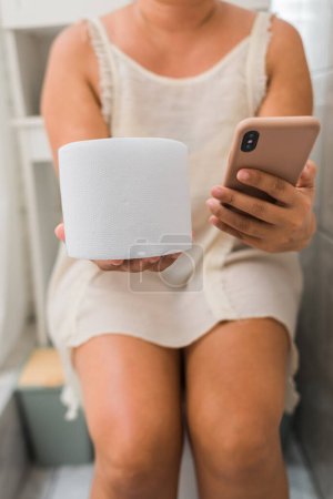 Téléchargez les photos : Femme tient son téléphone portable et son papier toilette dans ses mains alors qu'elle est assise sur les toilettes de sa maison. - en image libre de droit