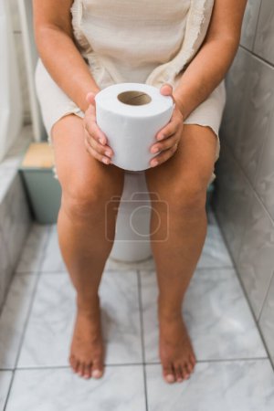 Mujer irreconocible sentada en el inodoro de su baño con papel higiénico