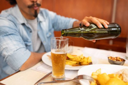 Photo for Unrecognizable man pours a golden beer from a green bottle into a glass sitting at the table with plates of Peruvian food. - Royalty Free Image