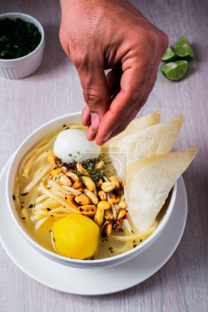 Foto de La mano de un latino vertiendo orégano sobre una sopa de pollo con wantan, fideos, huevo, patata amarilla, limón y cebolla china. - Imagen libre de derechos
