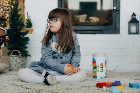 Téléchargez les photos : Une petite fille trisomique joue dans la maison près de la cheminée la veille de Noël. Une fille construit une tour en bois. - en image libre de droit