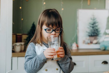 Téléchargez les photos : Une fille trisomique boit du jus d'orange et s'assoit à la table la veille de Noël. - en image libre de droit
