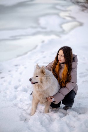 Photo for A girl walks with her beloved pet Samoyed in winter on the shore of a lake in the park. Walking the dog in winter. - Royalty Free Image
