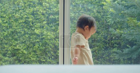 A small child in a cream outfit stands at a window looking out at a lush green garden, a moment of childhood curiosity and wonder captured against the backdrop of nature