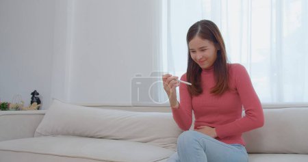 Ecstatic young asian woman in pink turtleneck holding looking at a pregnancy test and surprise exciting reaction laughing smile with happiness on a white couch, celebrating good news, a moment of joy