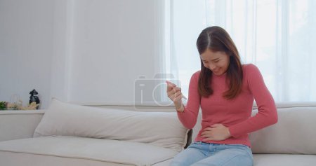 Téléchargez les photos : Jeune femme asiatique extatique au col roulé rose tenant en regardant un test de grossesse et réaction excitante surprise rire sourire avec bonheur sur un canapé blanc, célébrer de bonnes nouvelles, un moment de joie - en image libre de droit