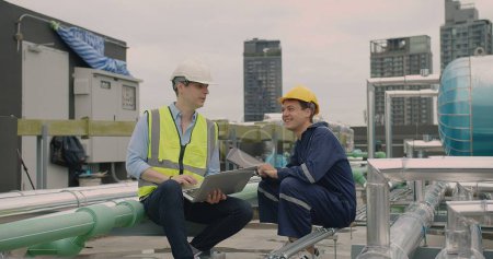 Zwei Ingenieure in Schutzhelmen und Westen arbeiten mit einem Laptop auf dem Dach, umgeben von Klimaanlagen