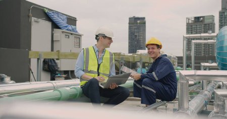 Zwei Ingenieure in Schutzhelmen und Westen arbeiten mit einem Laptop auf dem Dach, umgeben von Klimaanlagen