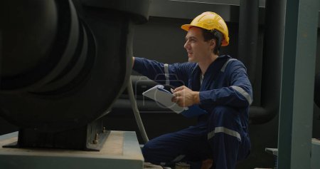 Industriearbeiter in blauem Overall und gelbem Bollenhut begutachtet aufmerksam Maschinen mit Klemmbrett in der Hand.