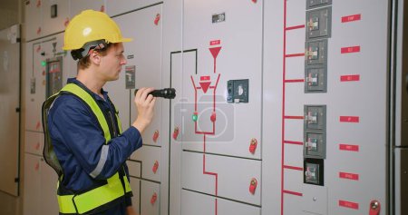 Techniker in Warnweste und gelbem Helm mit Taschenlampe, um Schaltschränke in einem Stromverteilerraum zu überprüfen