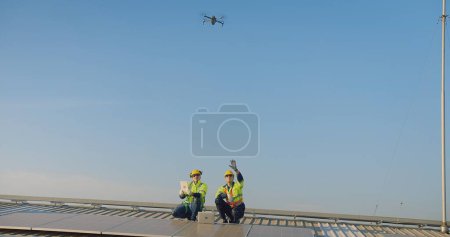 Two engineers with safety vests and hard hats are inspecting solar panels with a drone flying overhead against a clear blue sky
